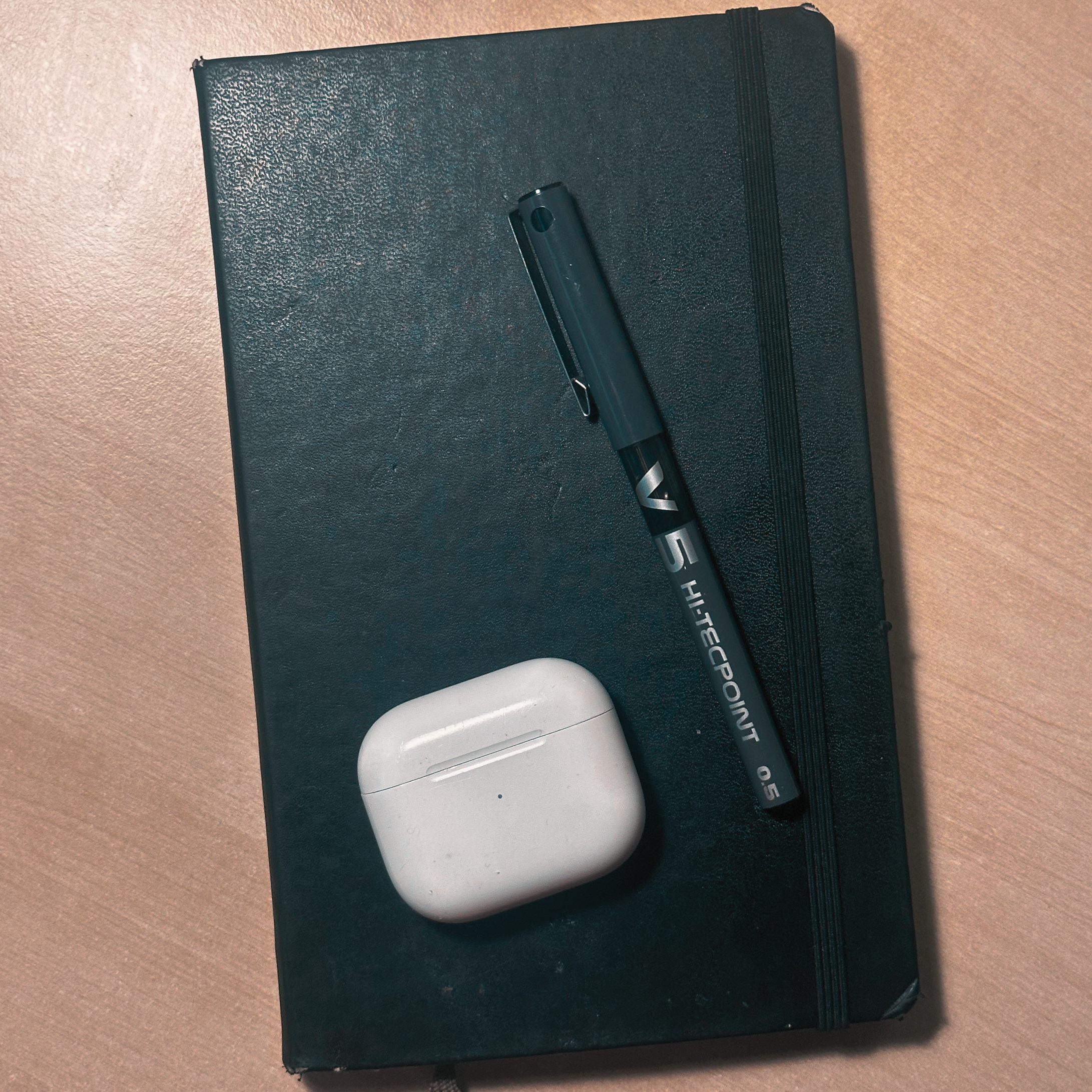 A black notebook with a pen and a pair of white wireless earbuds placed on top.